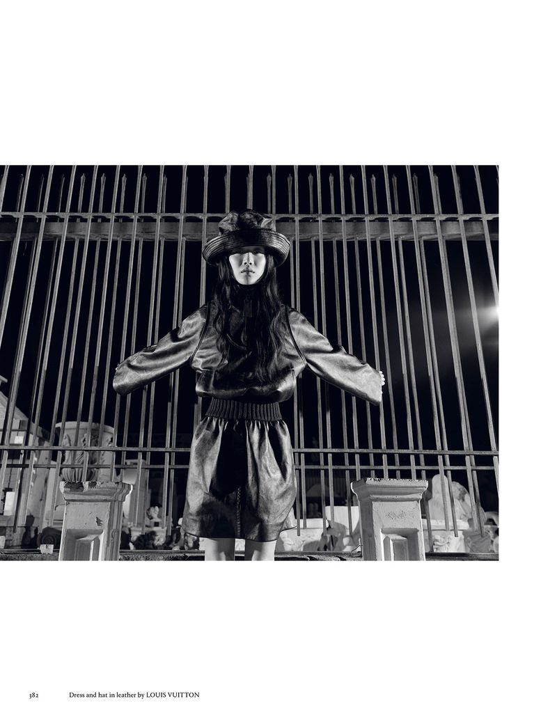 Fashion shot of a woman in a leather-look jacket and skirt standing in front of an iron gate at night, wearing a hat.
