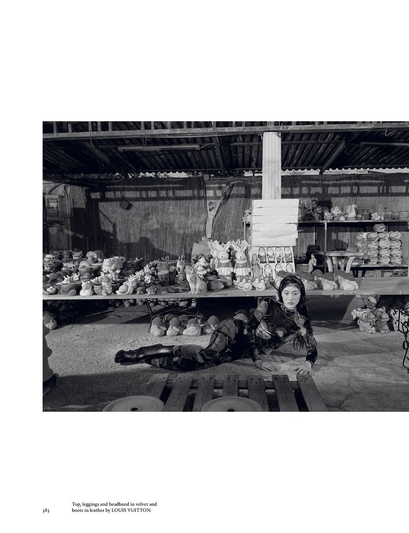 A woman in a dark patterned outfit lies on a wooden pallet indoors surrounded by shelves filled with ceramics and other items.