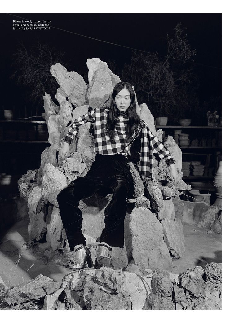 A woman in a checkered top and velvet pants sits on a rock structure in a dimly lit setting, with shelves of pottery visible in background.