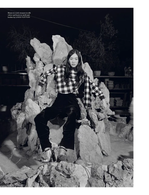 A woman in a checkered top and velvet pants sits on a rock structure in a dimly lit setting, with shelves of pottery visible in background.