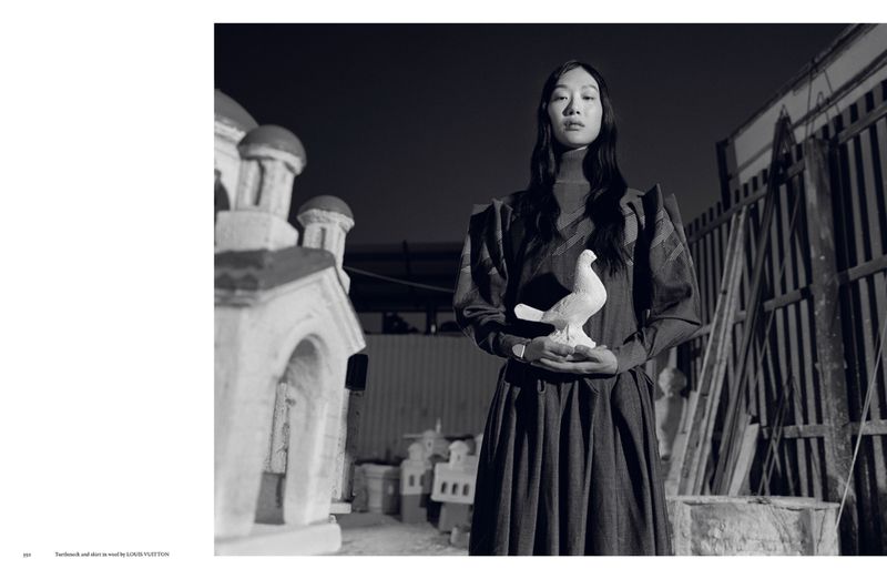 Monochrome studio shot of an Asian woman in dark dress holding a ceramic dove, with building props visible in the background.