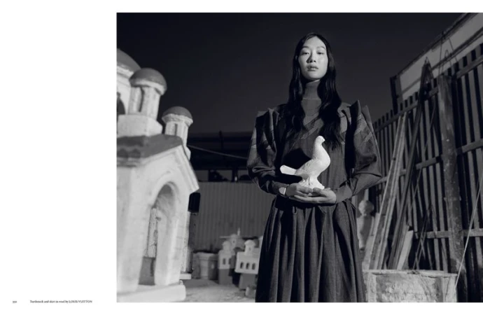 Monochrome studio shot of an Asian woman in dark dress holding a ceramic dove, with building props visible in the background.