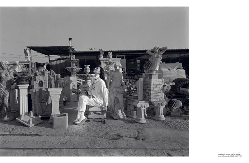 Monochrome shot of an adult female in white sitting amongst various concrete statues and outdoor decor under a bright sky, in daylight.