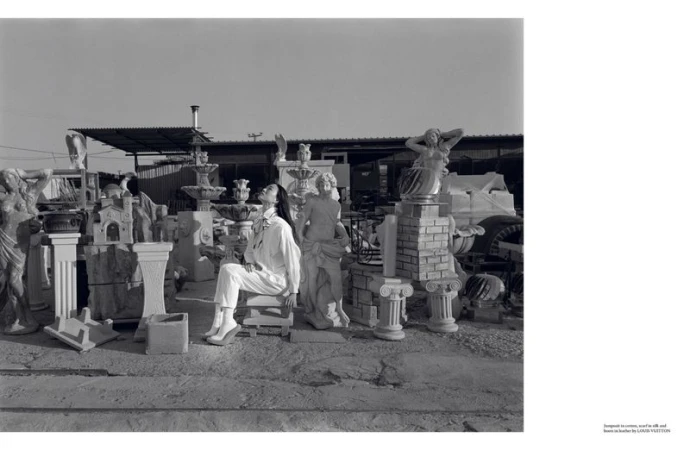 Monochrome shot of an adult female in white sitting amongst various concrete statues and outdoor decor under a bright sky, in daylight.