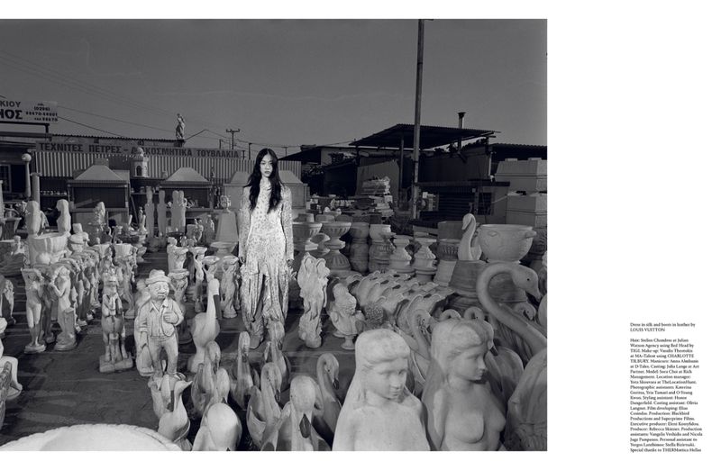 Fashion shot of a slender woman in a long dress, standing in a yard full of concrete statues and decorative outdoor pottery.