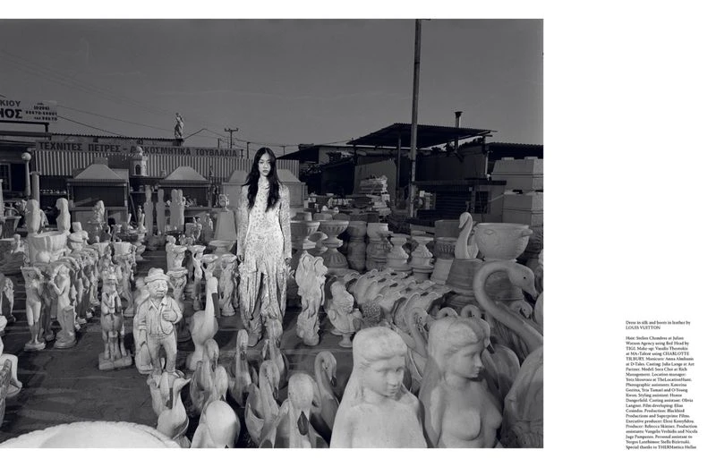 Fashion shot of a slender woman in a long dress, standing in a yard full of concrete statues and decorative outdoor pottery.
