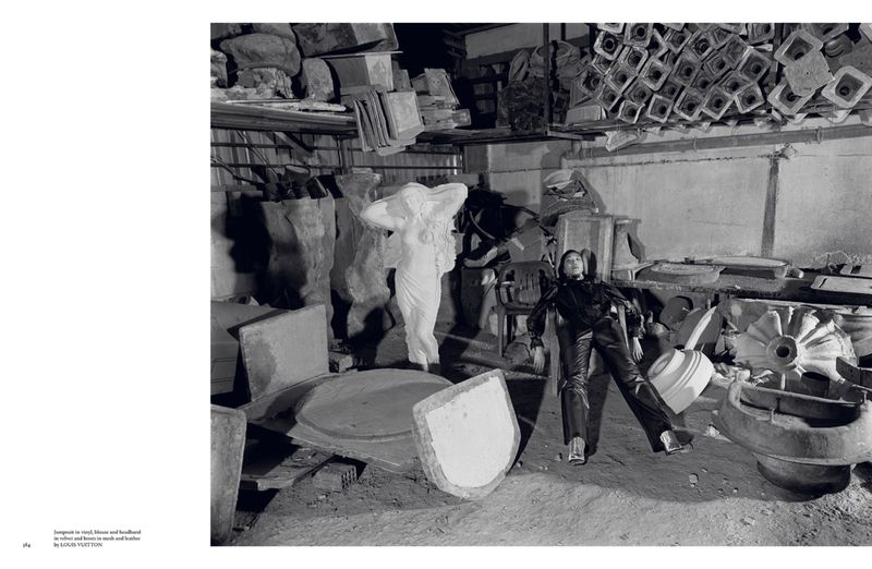 A young woman in leather attire poses among discarded concrete forms in a weathered indoor industrial setting, captured in black and white.