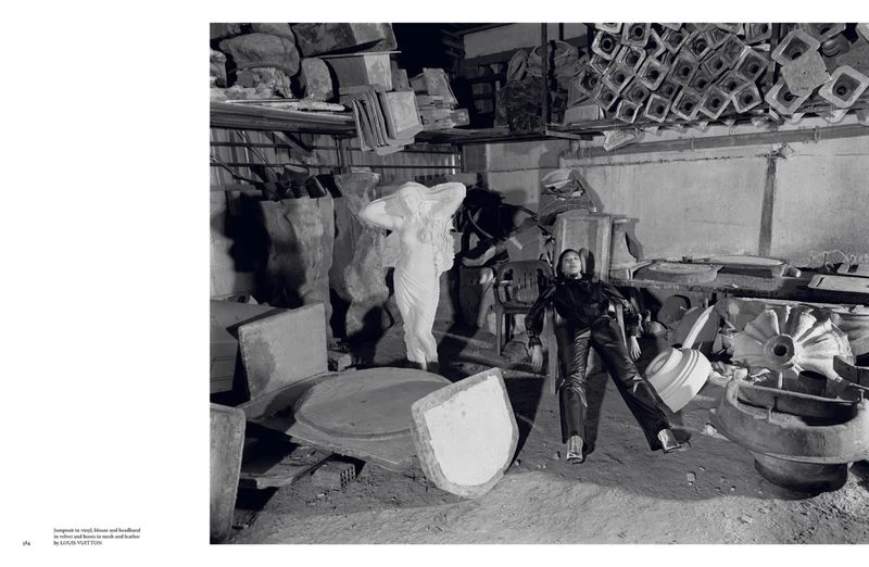A young woman in leather attire poses among discarded concrete forms in a weathered indoor industrial setting, captured in black and white.