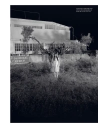 Model Sora Choi stands in an overgrown field at night wearing a vinyl trench coat before an industrial building.