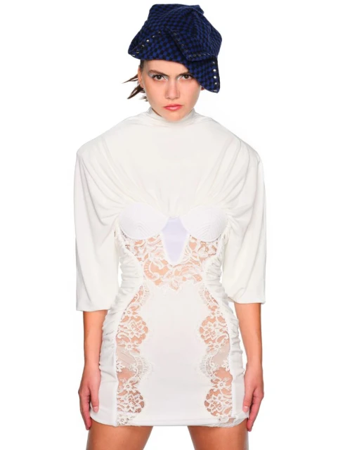 Fashion model poses in a white mini dress with lace detail and a blue cap with silver accents against a stark white background.