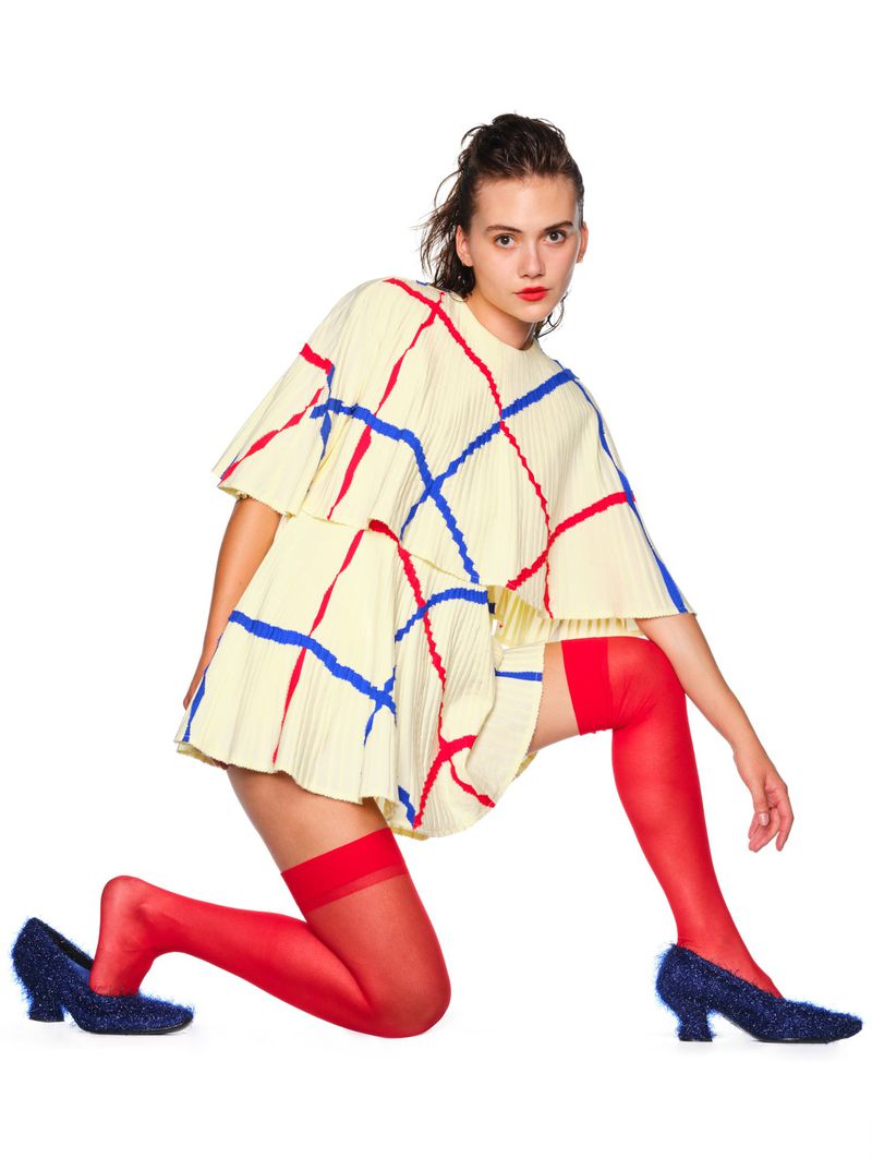 A woman in a patterned, yellow two-piece outfit poses kneeling against a white backdrop, wearing red stockings and sparkly blue heels.