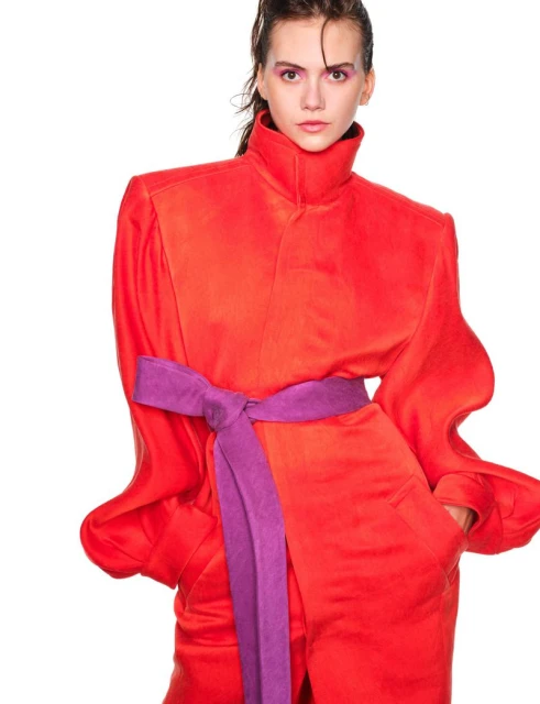 A fashion model in a bold red, high-necked jumpsuit with a purple sash, poses against a white backdrop in a studio setting.