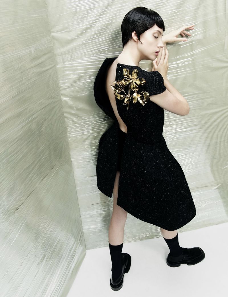 A short-haired woman poses in a black dress with a gold floral accent against a textured white background, wearing black socks and platform shoes.
