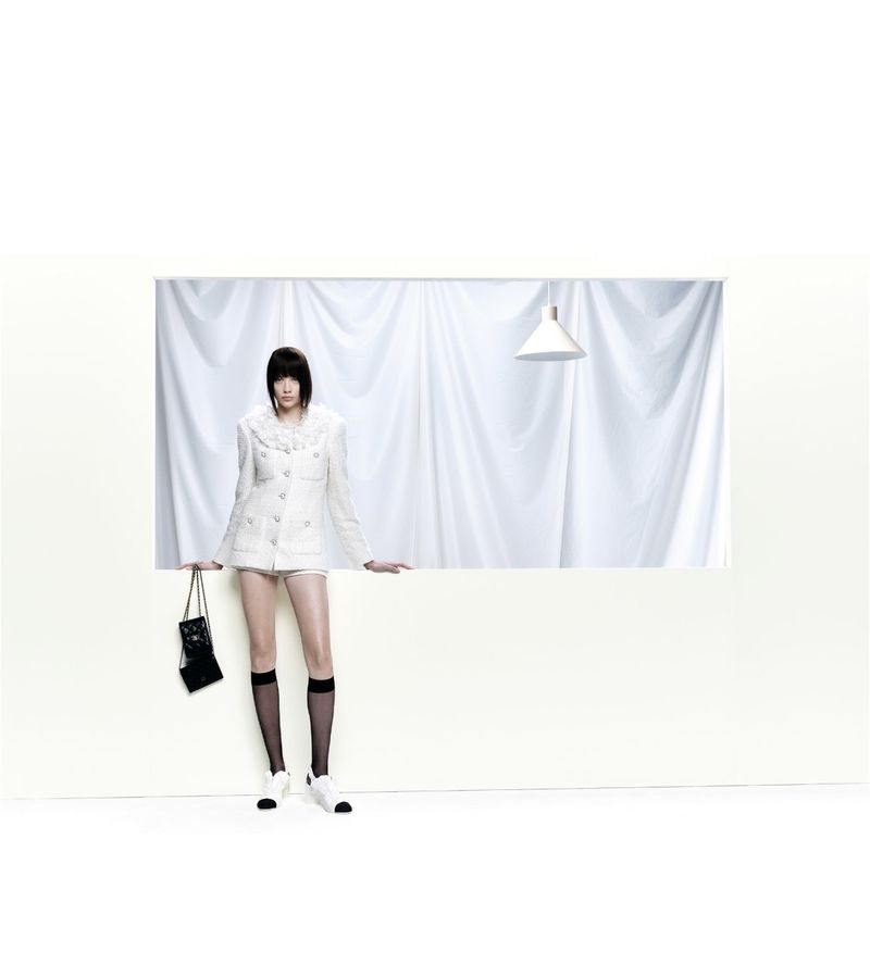 A stylishly dressed young woman in a white jacket, shorts, and knee-high socks poses in a minimalist indoor setting with a handbag.