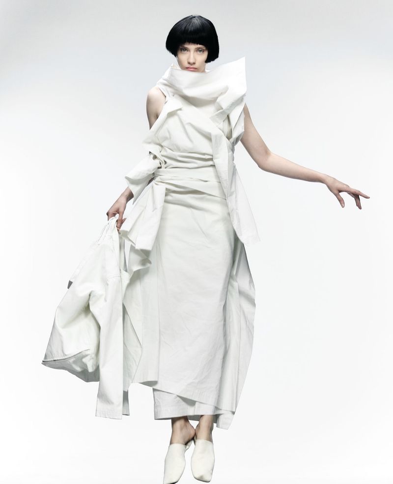 An adult female model wearing a voluminous white dress and white shoes, posing against a white studio background. Fashion editorial.