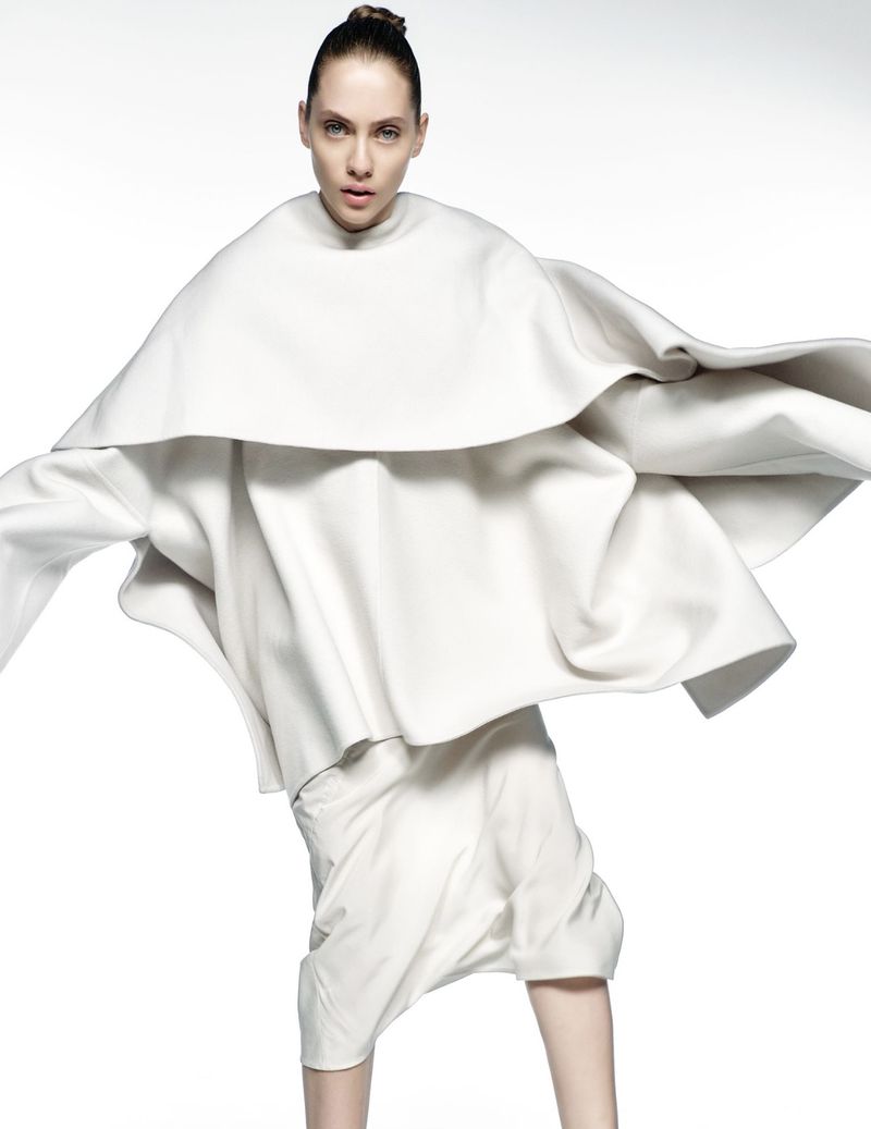 Woman in white sculptural layered dress on white background in studio. Fashion shot with neutral expression and minimal hair style.
