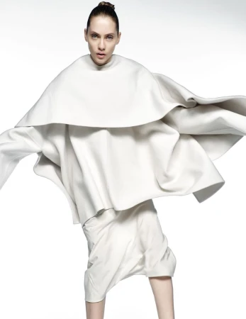 Woman in white sculptural layered dress on white background in studio. Fashion shot with neutral expression and minimal hair style.