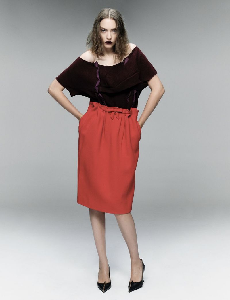 Fashion model poses in maroon top and red skirt with black heels against a gray background in a studio shot, looking at camera.
