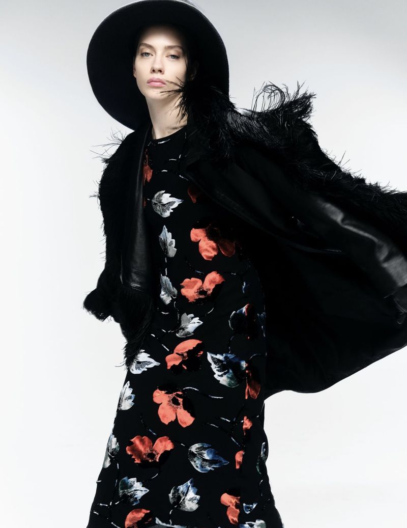 Odette Pavlova wearing a wide-brim black hat and floral dress with a billowing feathered coat against a white studio background.
