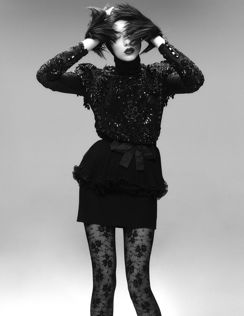 Monochrome fashion portrait of a woman in a black beaded top, mini skirt, and patterned tights, posing with hands near her head.