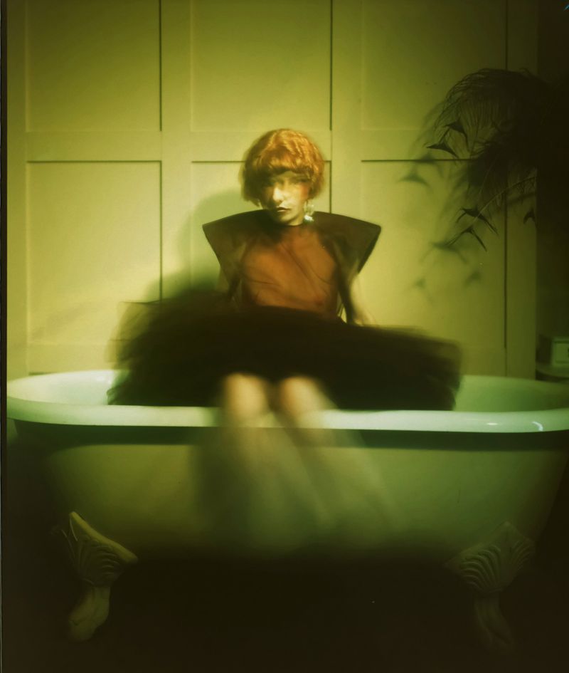 A young person with red hair sits on the edge of a vintage-style bathtub, wearing a unique orange top and black tulle skirt, inside a room.