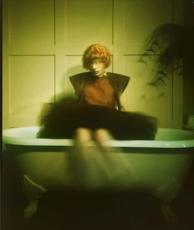 A young person with red hair sits on the edge of a vintage-style bathtub, wearing a unique orange top and black tulle skirt, inside a room.