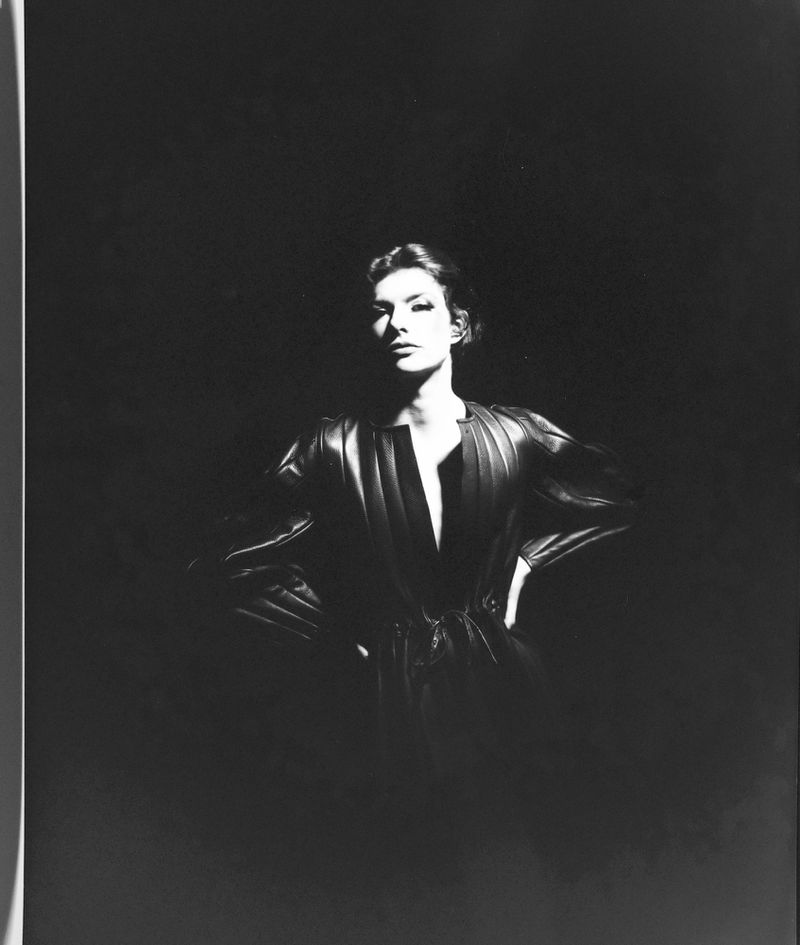 Monochrome portrait of a woman with short hair wearing a shiny, dark outfit with hands on hips in a dimly lit studio setting.