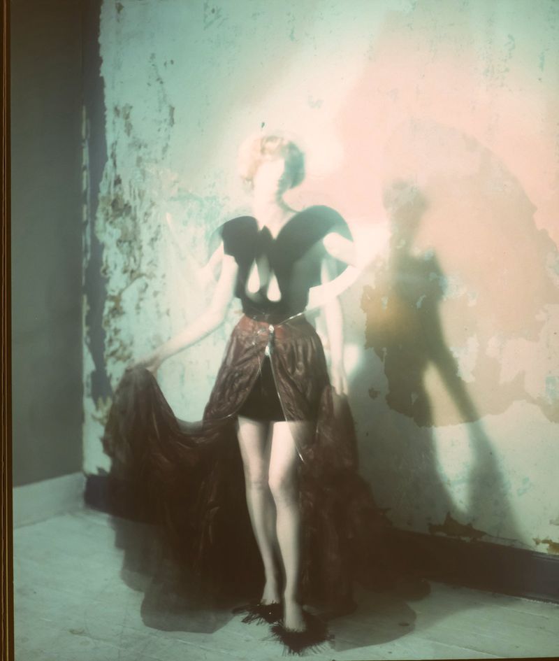 An adult female poses indoors wearing a distinctive fashion outfit and dark shoes with a feathered texture, set against a textured wall.