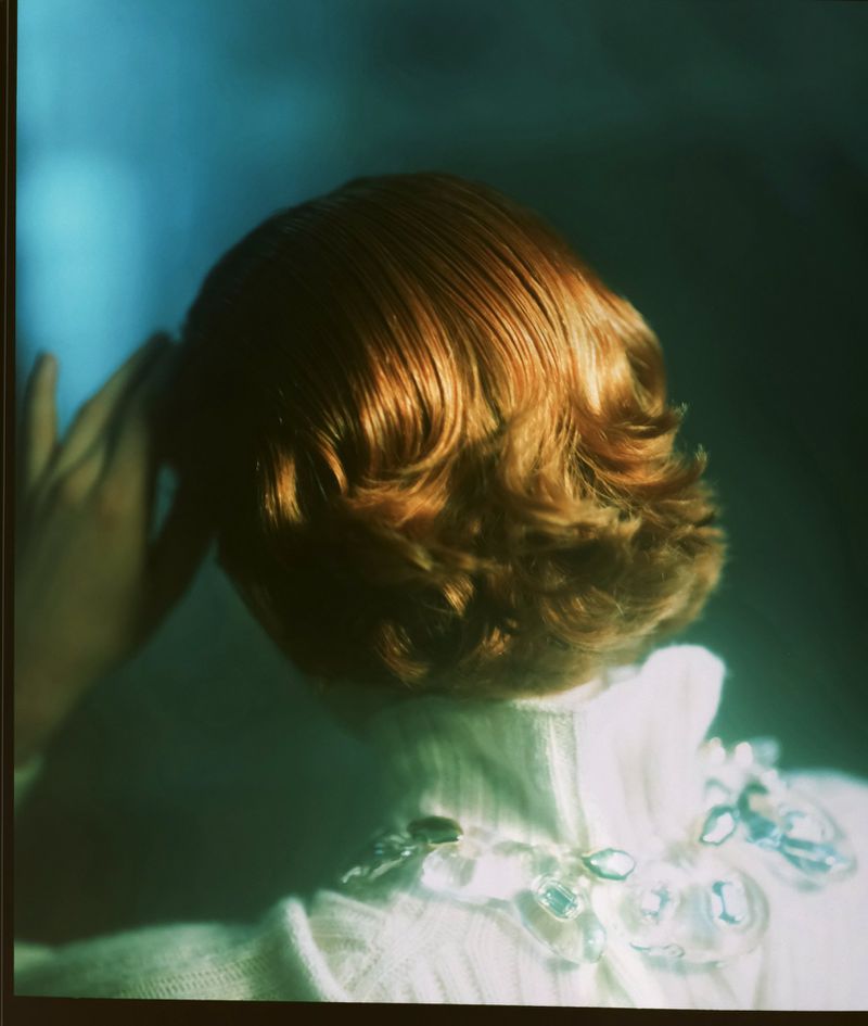 Back view of a woman with short, wavy reddish-brown hair wearing a white collared shirt with clear jewelry against a teal background.