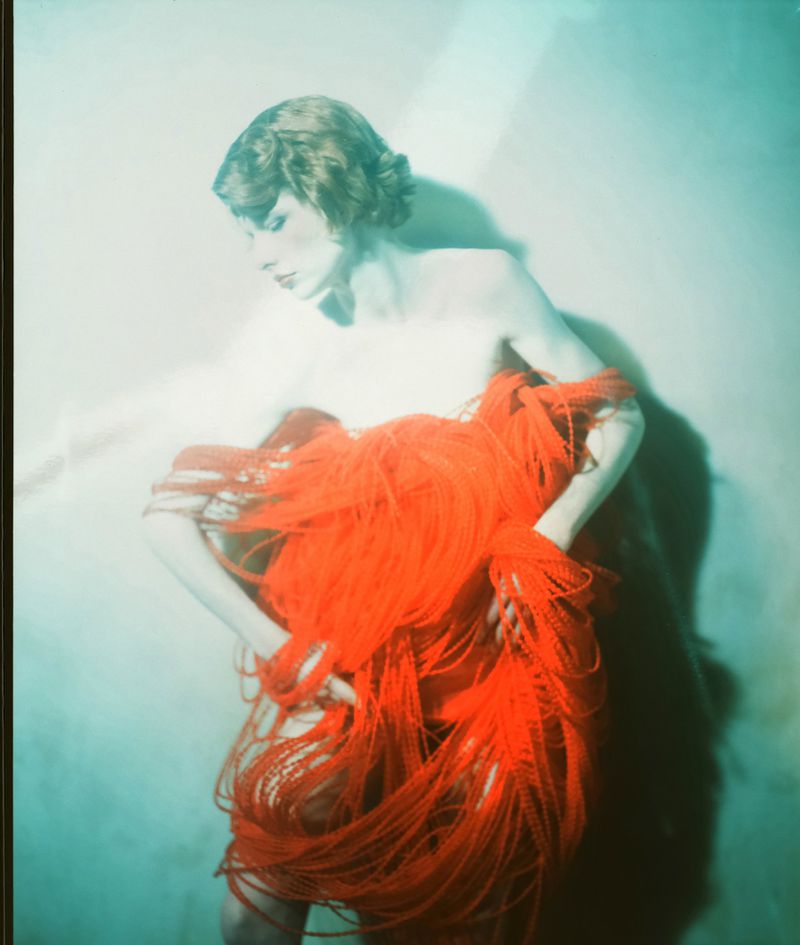 Woman with a red string garment in a turquoise studio setting. The scene features diffused lighting and a moody atmosphere.