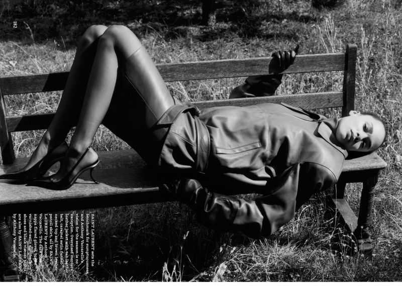 Monochrome fashion shot of a model in leather jacket and skirt lounging on a wooden bench in a grassy field, wearing heels and stockings.