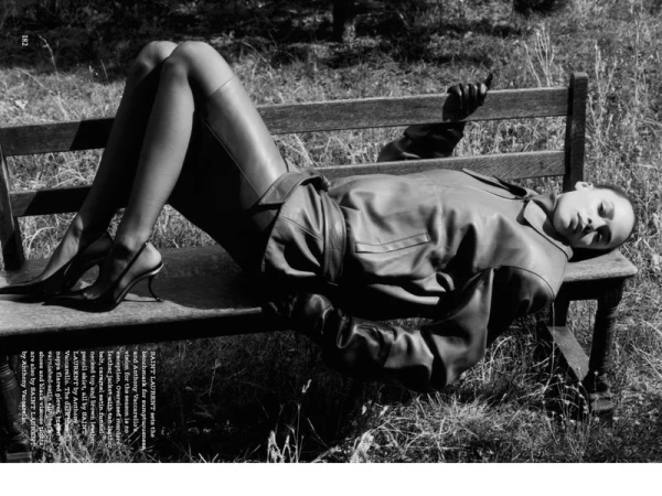 Monochrome fashion shot of a model in leather jacket and skirt lounging on a wooden bench in a grassy field, wearing heels and stockings.