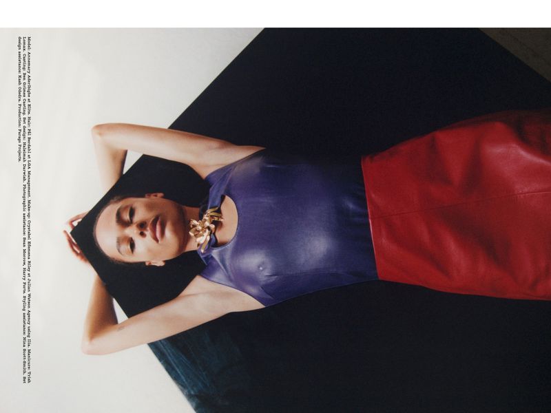Woman in a blue top and red skirt is lying on a black cushion. She is wearing gold necklace and has her arms above her head.