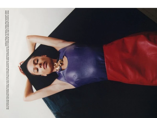 Woman in a blue top and red skirt is lying on a black cushion. She is wearing gold necklace and has her arms above her head.