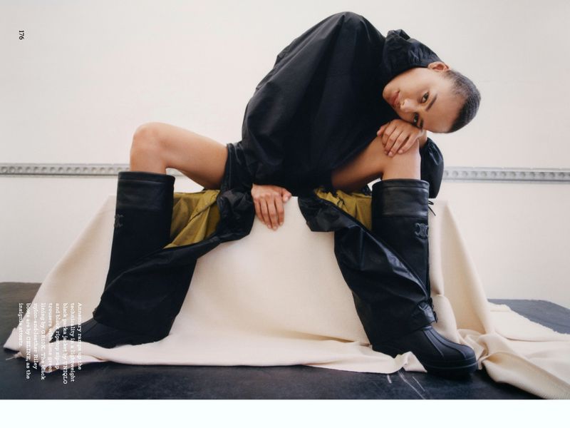 Fashion shot of a model in a black hooded jacket and black boots sitting on a draped surface in a studio with a white background.