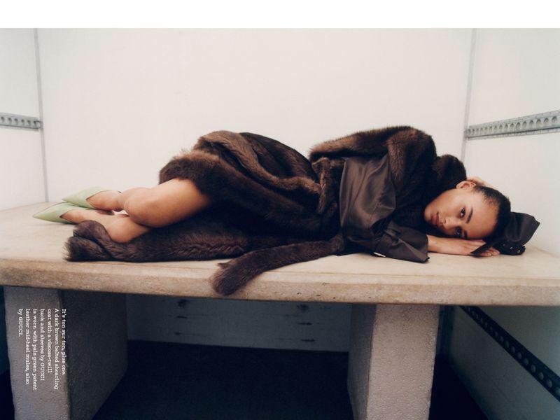 Adult female lying on a stone table wearing a brown shearling coat and green heels in a minimalist studio environment.