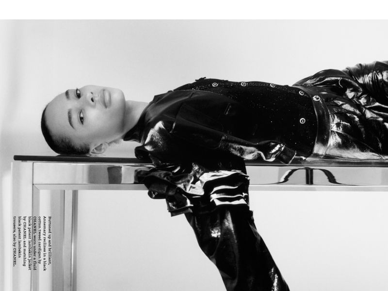 Annemary reclines in a black cotton tweed cardigan and black patent lambskin jacket and trousers, all by Chanel, in a studio shot.