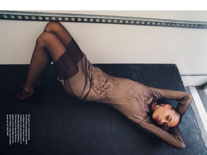 Fashion editorial image: A woman reclines in a sheer brown dress and black tights on a dark surface with a minimalist background.