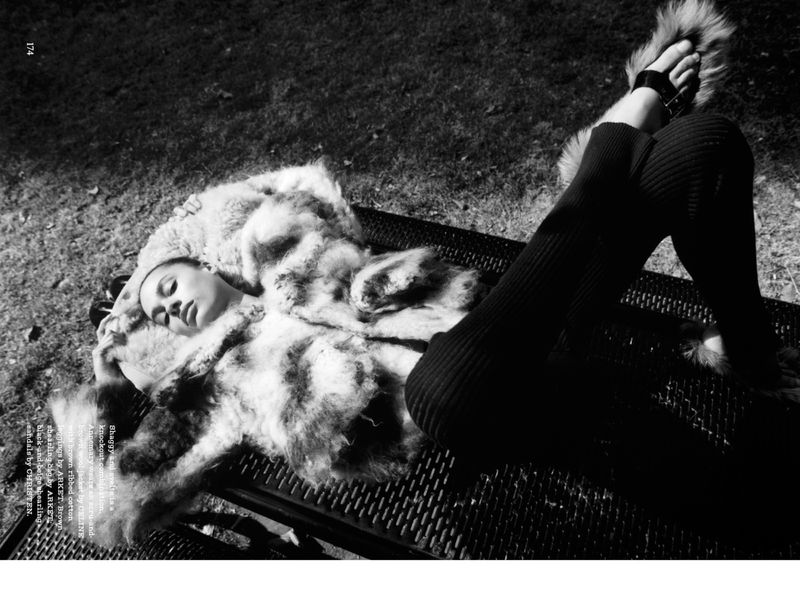 Adult woman lies on a metal bench outdoors, resting on a fur blanket, wearing fur shoes, shot in black and white, under diffused sunlight.