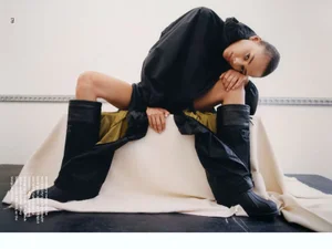 Model Annemary Aderibigbe wearing an oversized black parka jacket and tall black boots posing dynamically on a draped white bench.