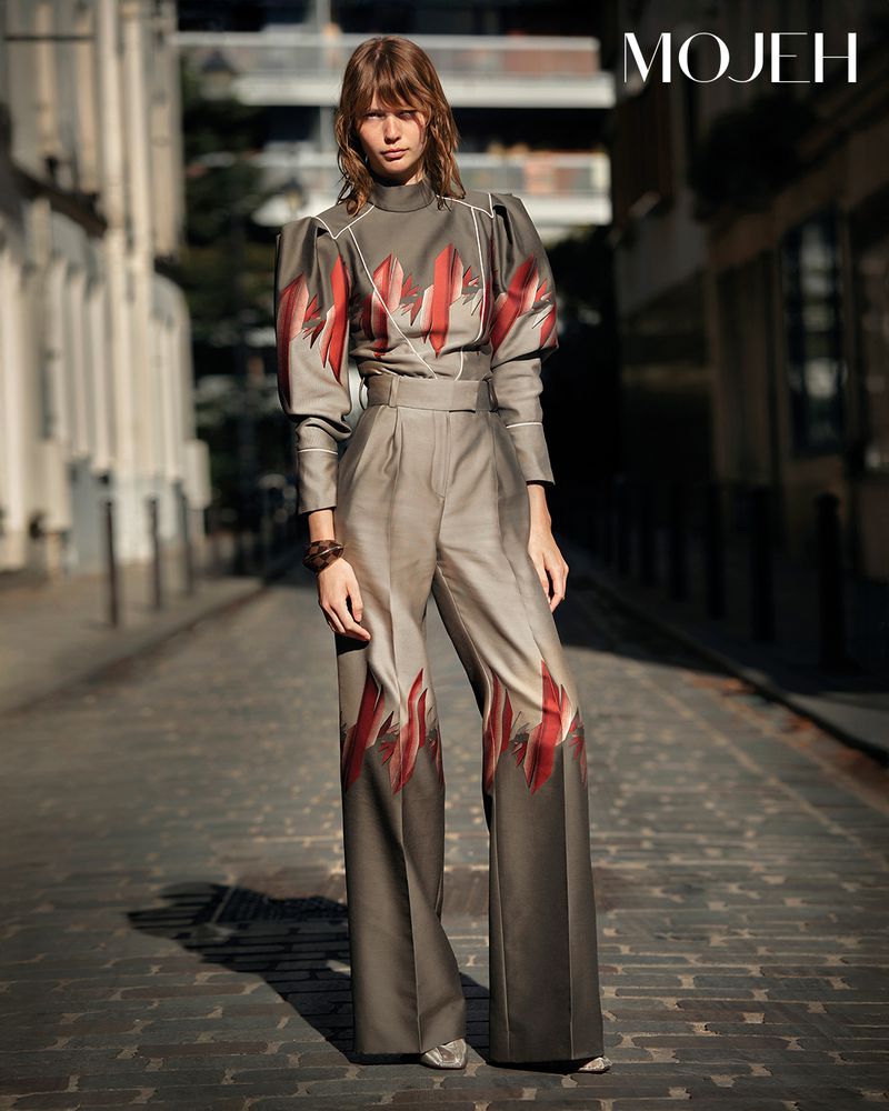 Fashion model in a gray two-piece suit with red accents poses on a cobblestone street in an urban setting with MOJEH logo.