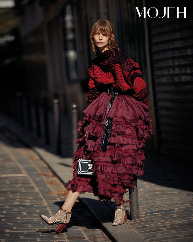 Fashion model poses in a red and black sweater with a burgundy, tiered, ruffled skirt on a city street in an editorial shot.