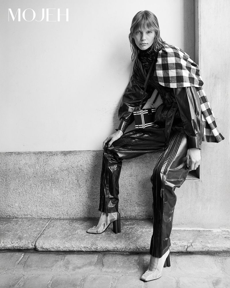 A single fashion model poses indoors wearing a shiny black vinyl outfit with transparent heels, a plaid wrap, and a small handbag.