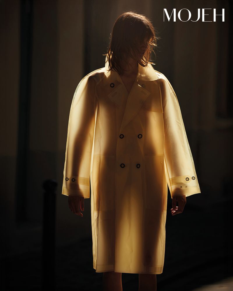 Fashion shot featuring an androgynous model wearing a transparent yellow trench coat in a dimly lit, modern outdoor setting.