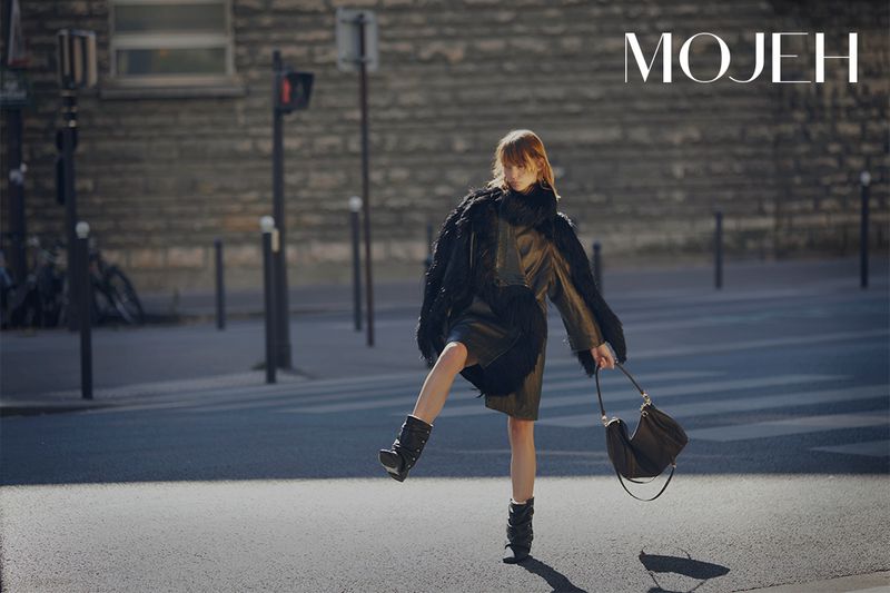 A woman in a black dress and boots strides confidently on a city street in a fashion editorial shot, carrying a stylish handbag.