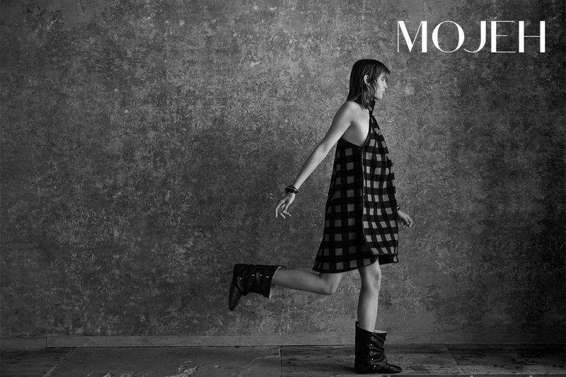 Black-and-white fashion shot of a woman in a patterned dress and boots, walking against a textured wall, with 'MOJEH' logo.