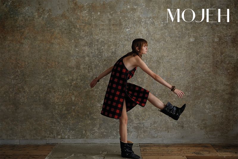 A woman in a red plaid dress with black boots strikes a playful pose in front of a textured neutral backdrop with the MOJEH logo.