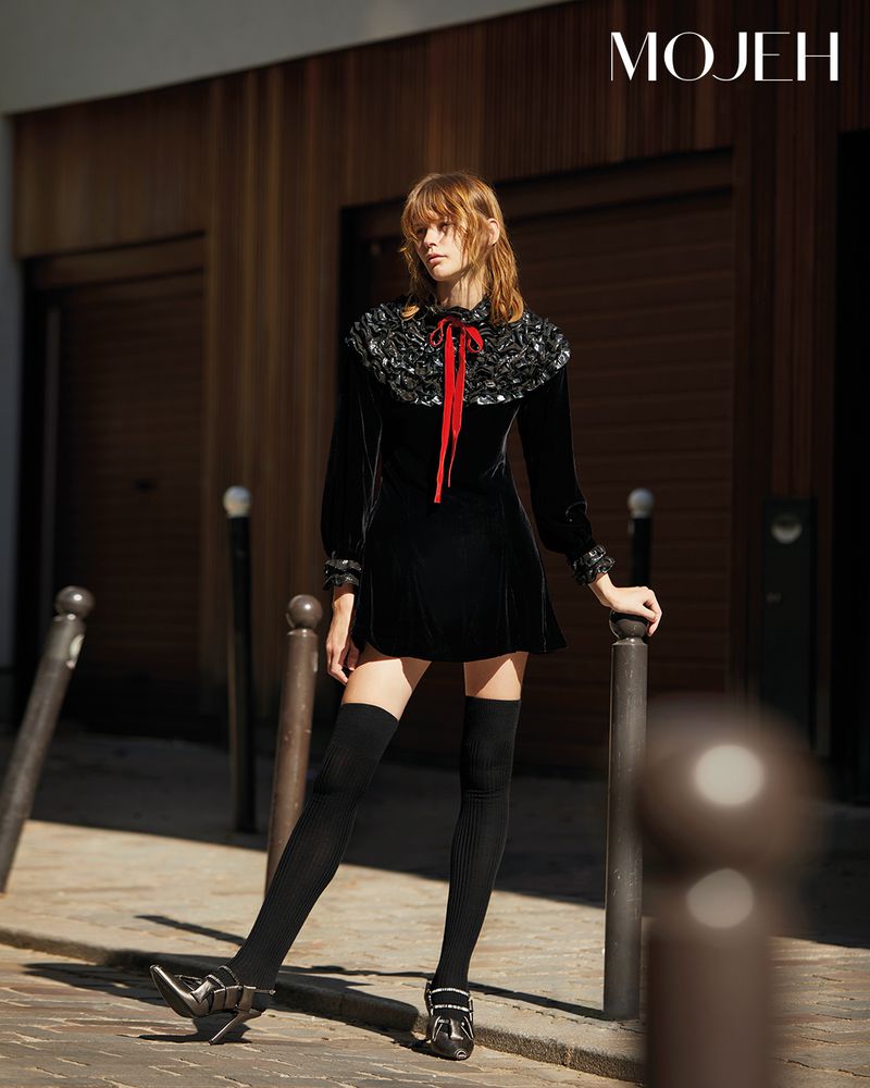 Elegant woman poses on a city street in a black dress with long sleeves, dark stockings, and buckle shoes, standing near bollards.