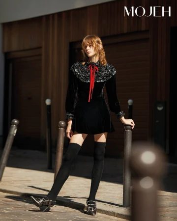 Elegant woman poses on a city street in a black dress with long sleeves, dark stockings, and buckle shoes, standing near bollards.