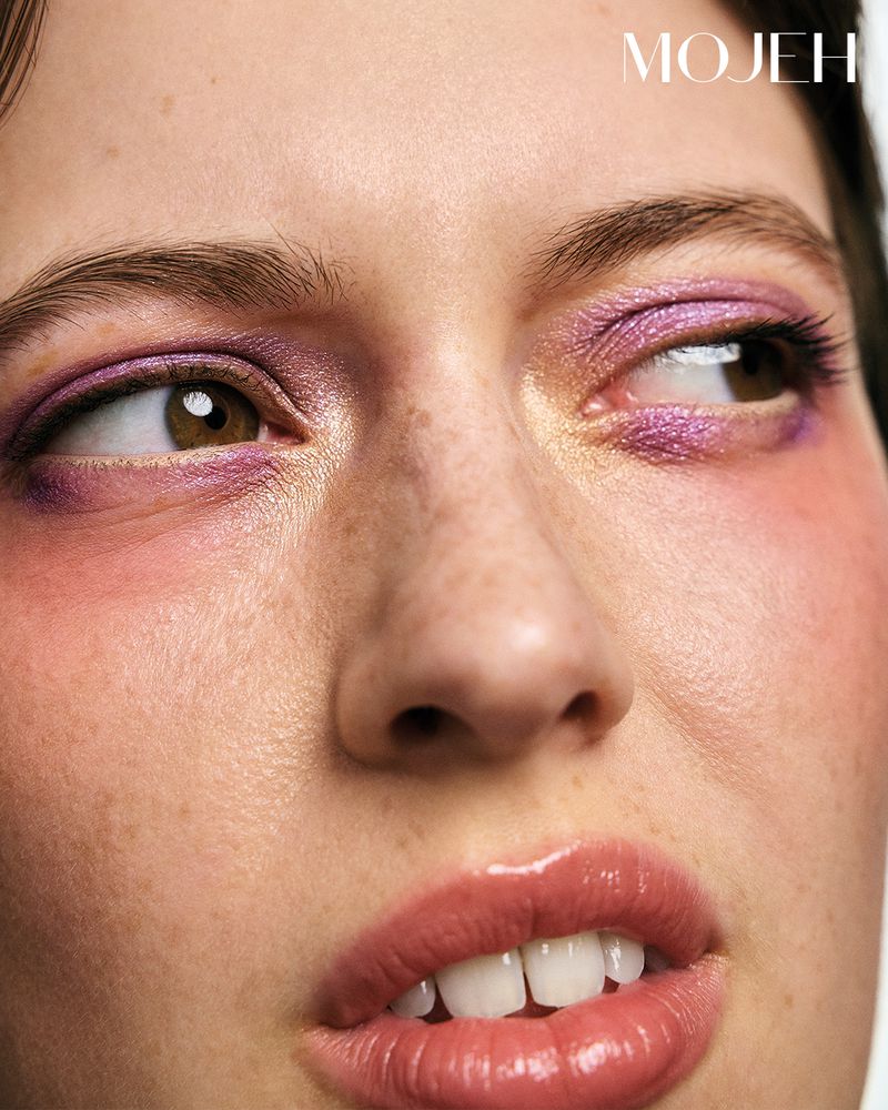 Close-up shot of a woman's face with glossy lips and purple eyeshadow. The image showcases makeup and skincare details in studio lighting.